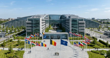 NAVO-hoofdkwartier in Brussel op een zonnige dag. Op de voorgrond wapperen de vlaggen van de lidstaten en de NAVO-vlag op een groot plein. Op de achtergrond is het moderne, glazen gebouw met zijn karakteristieke architectuur te zien, terwijl enkele afgevaardigden over het terrein lopen