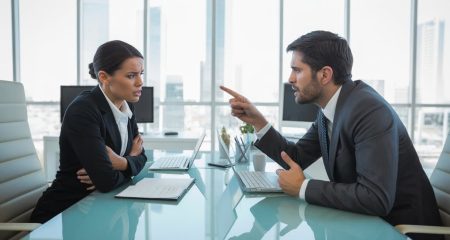 Twee kantoormedewerkers in een gespannen gesprek tegenover elkaar aan een bureau in een modern kantoor.