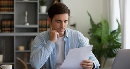 Een jonge man zit rustig aan een bureau en leest geconcentreerd een document in een thuiskantoor.