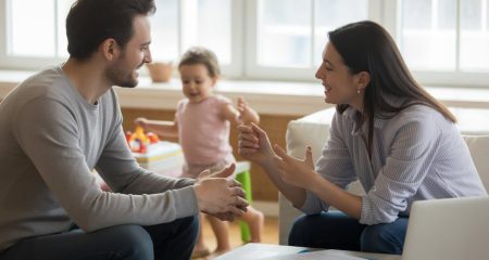 Twee volwassenen zitten samen aan een tafel met documenten, terwijl een kind op de achtergrond speelt in een lichte woonkamer.