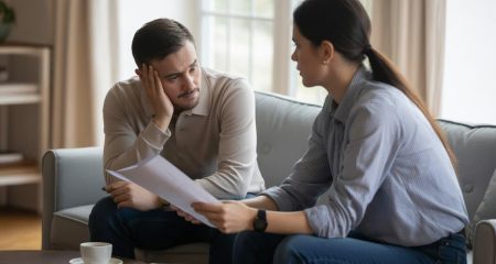 Twee volwassenen voeren een serieus gesprek in een woonkamer, één houdt een document vast en de ander luistert aandachtig.