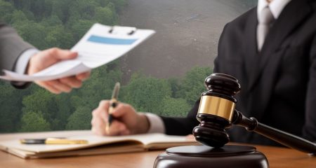 Een rechtershamer op een houten bureau met op de achtergrond een vervuilde natuur en handen die documenten uitwisselen.